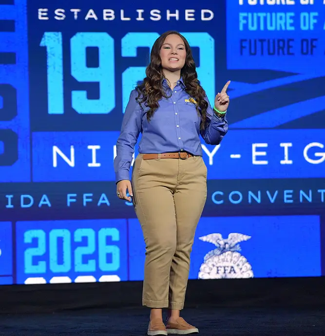 A woman FFA member in a blue shirt standing on stage, smiling and gesturing with one hand.