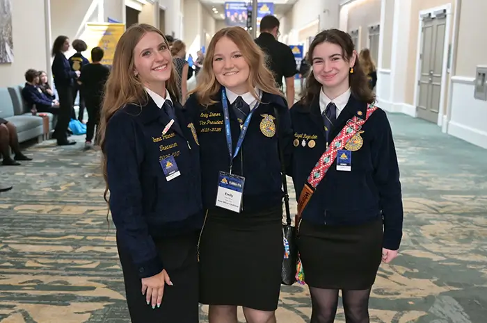 Three young women in official blue FFA jackets and black skirts stand together smiling in a convention hallway.